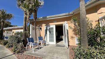 A building with a beige exterior, numbered 603, featuring a patio with two blue chairs and a small table, surrounded by palm trees and shrubs.