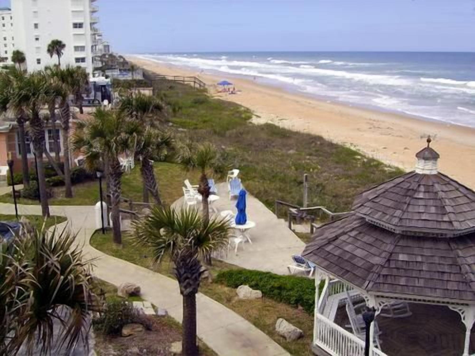 Vista de la playa con un pabellón, palmeras y un camino que conduce a la arena.