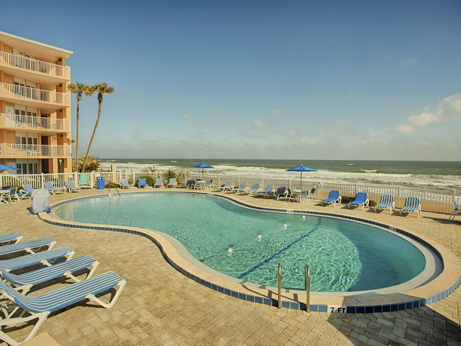 A swimming pool with lounge chairs and umbrellas, a beach view, and a building with balconies.