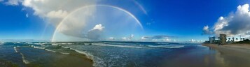 A beach with waves, a rainbow, and a building in the distance.