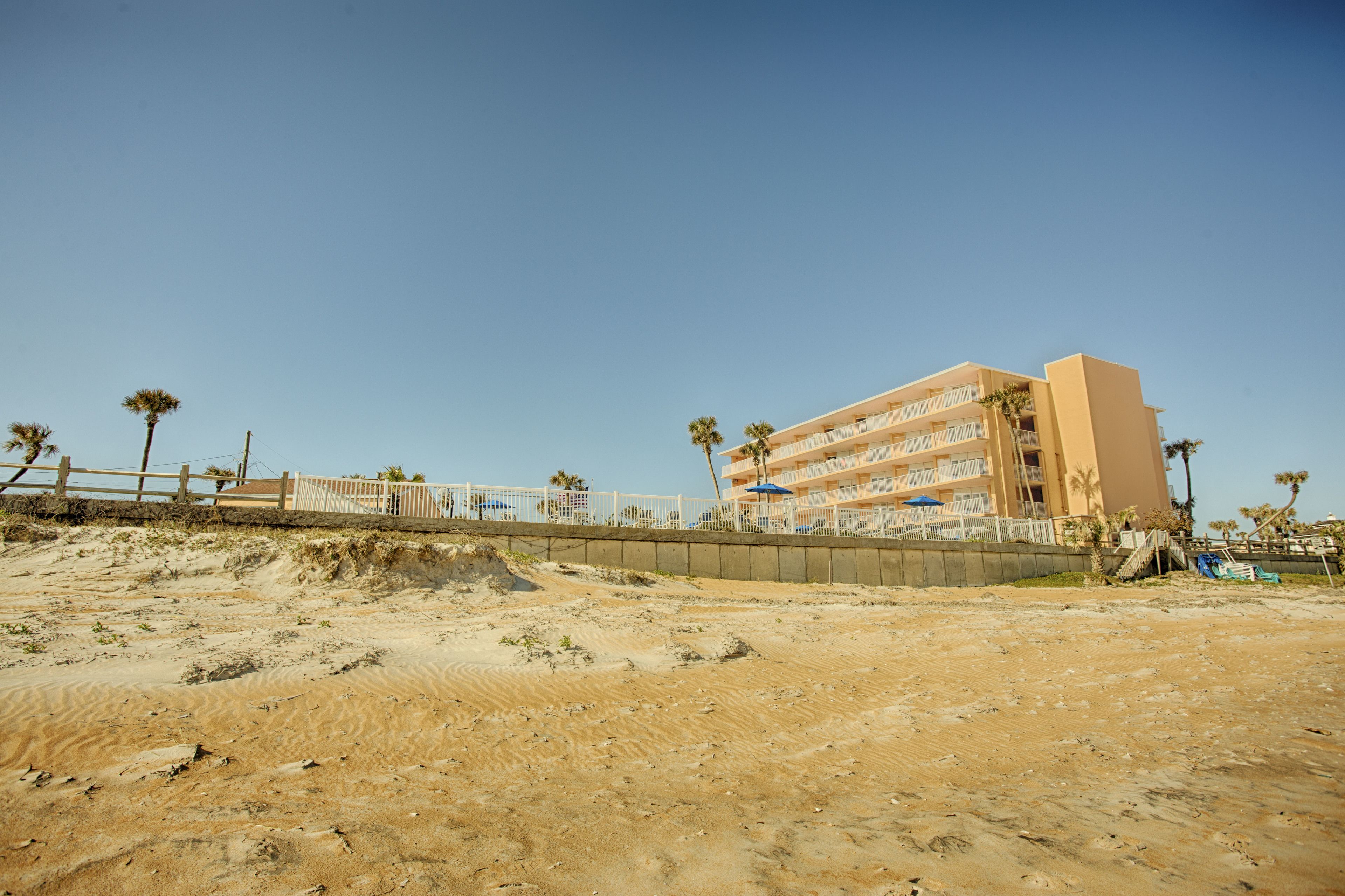 A beachfront hotel with multiple floors, a sandy beach in the foreground, and palm trees.
