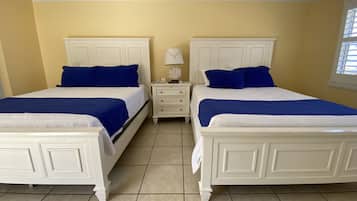 Quarto duplo com duas camas, cada uma com cabeceira branca e roupa de cama azul, mesa de cabeceira com abajur e janela com persianas.
