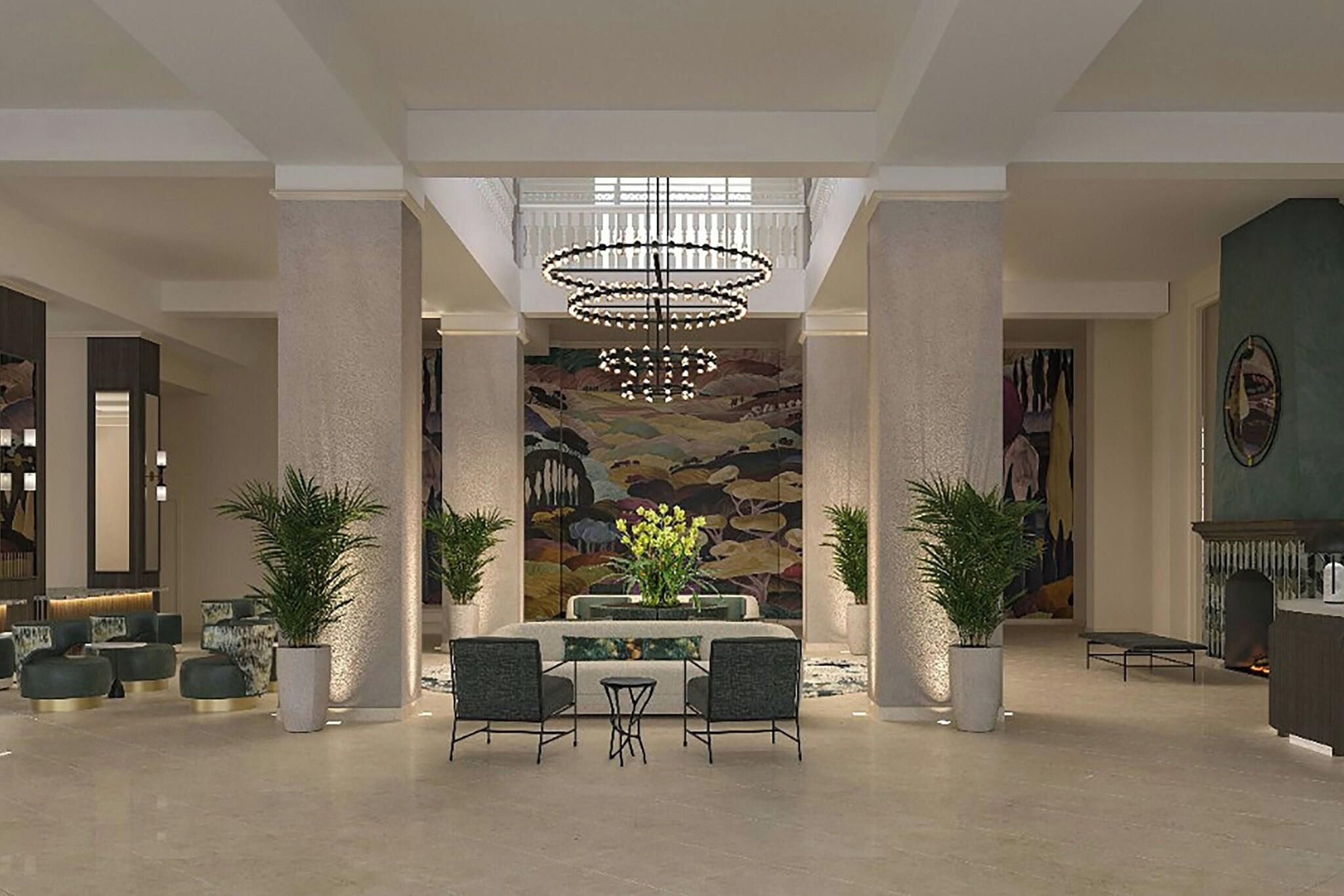 A spacious lobby with a high ceiling, a central chandelier, a dining area with a table and chairs, and a large mural on the back wall.