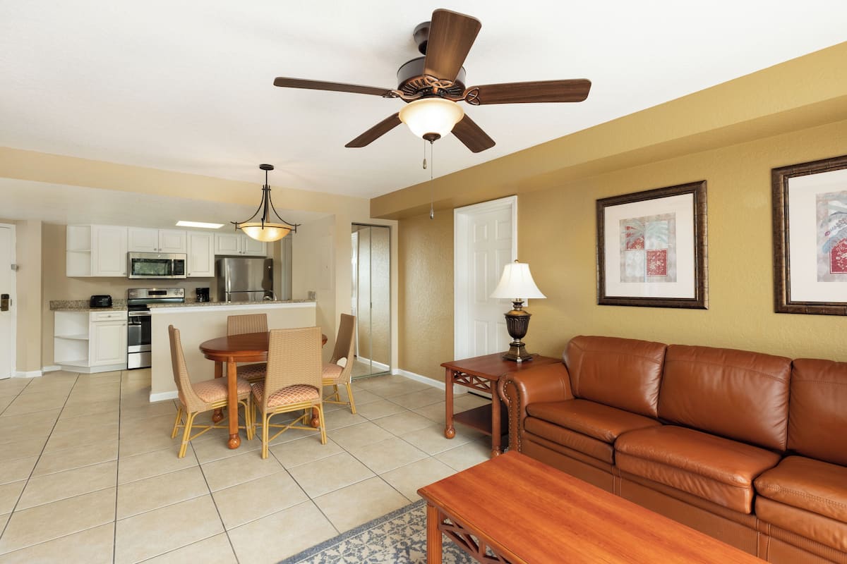 A modern kitchen with white cabinets, a stainless steel refrigerator, and a microwave. There is a dining area with a wooden table and wicker chairs. A brown leather sofa and a small wooden table with a lamp are in the living area.