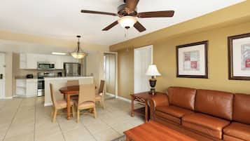 A modern kitchen with white cabinets, a stainless steel refrigerator, and a microwave. There is a dining area with a wooden table and wicker chairs. A brown leather sofa and a small wooden table with a lamp are in the living area.