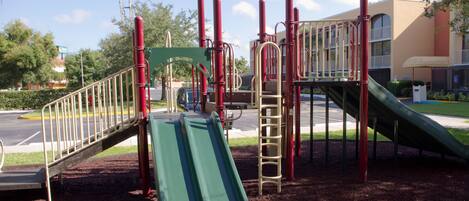 Un parque infantil con toboganes y estructuras para escalar, rodeado de un estacionamiento y edificios.