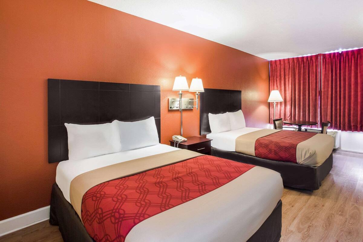 Ein Hotelzimmer mit zwei Betten, einer rot-beigen Tagesdecke, einem schwarzen Kopfteil, einem Nachttisch mit Lampe und einem Fenster mit roten Vorhängen.