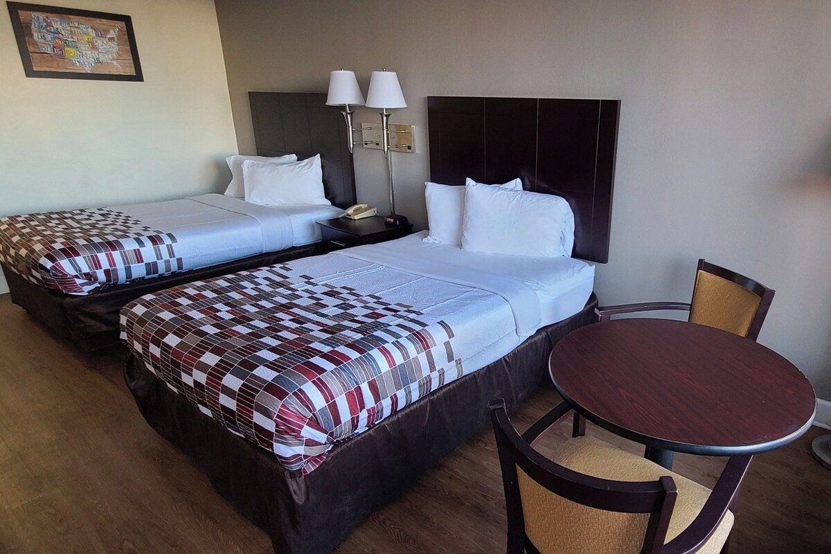 Quarto de hotel com duas camas, uma mesinha de cabeceira com luminárias, uma mesa de madeira redonda e uma cadeira.