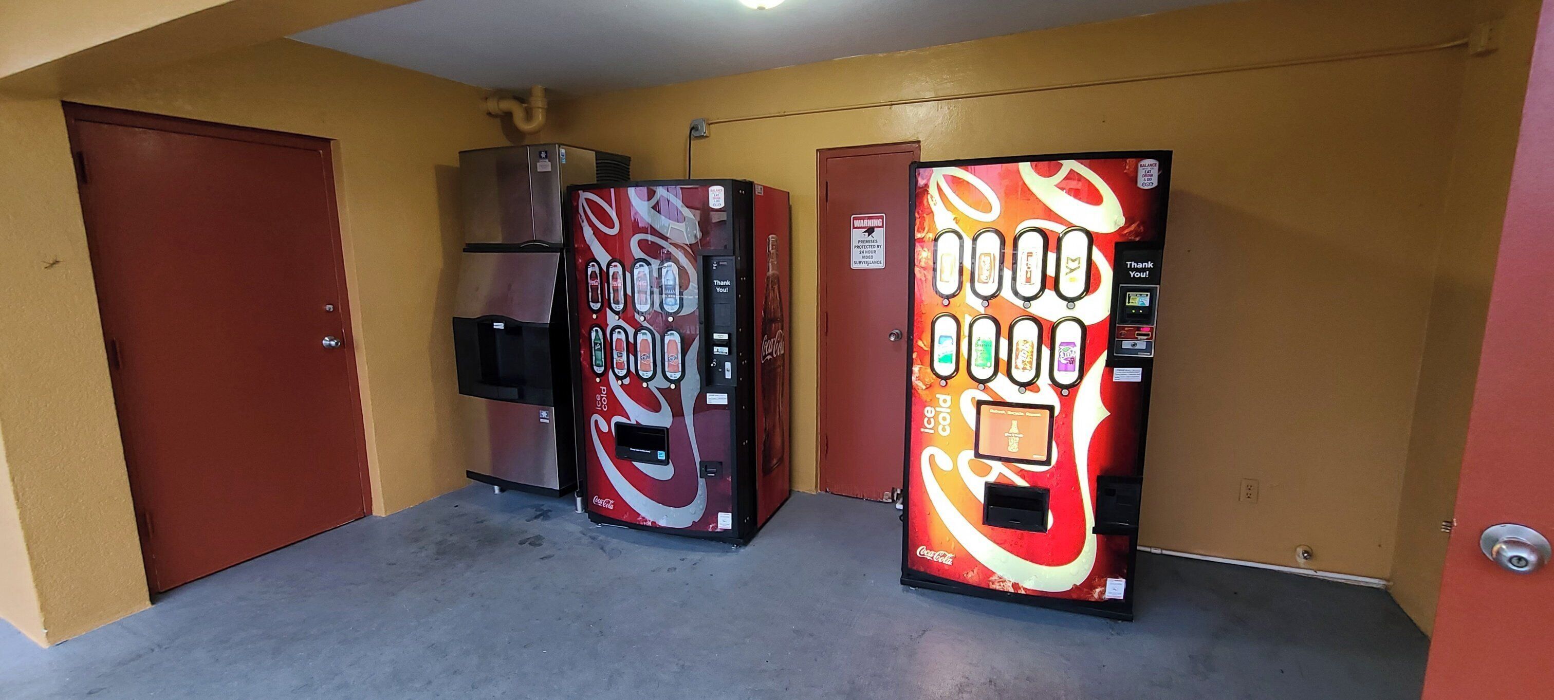Vending machines with Coca-Cola branding in a hallway with doors and a ceiling vent.