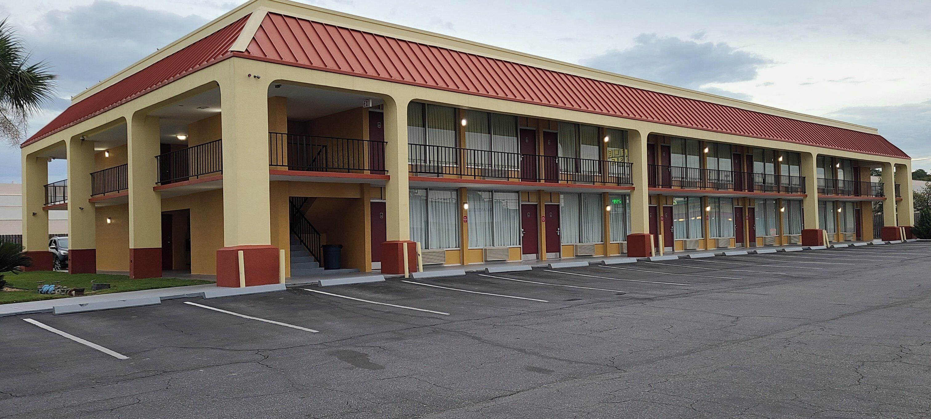 A two-story motel with a red roof and beige walls, featuring a central entrance with a staircase, multiple balconies, and a parking lot.