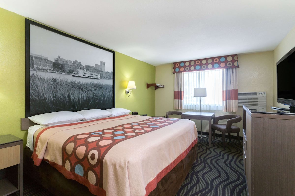 A hotel room with a bed, desk, chair, TV, and a large cityscape painting on the wall.