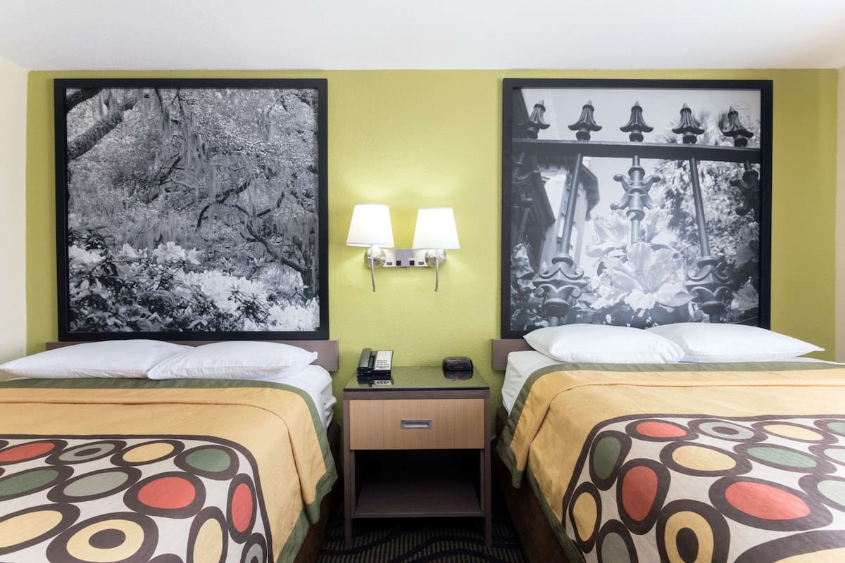 A hotel room with two beds, a nightstand with a lamp, and a wall-mounted phone.
