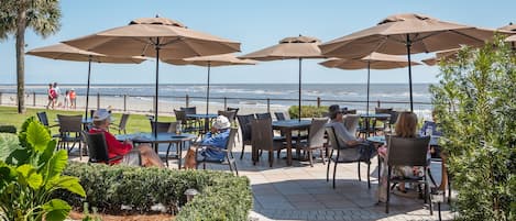 An outdoor patio with tables and chairs under umbrellas, overlooking the beach.