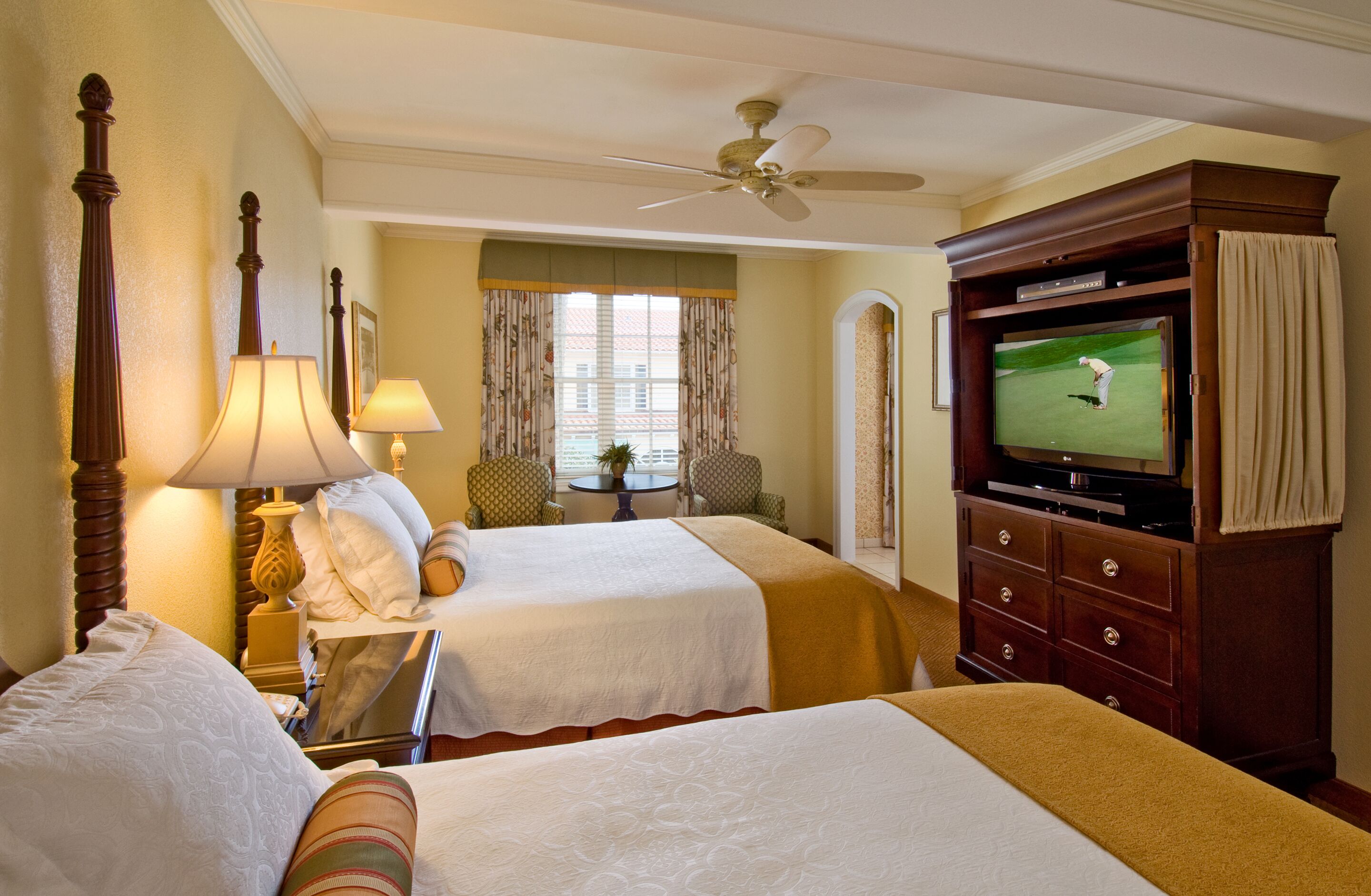 A hotel room with two beds, a television, a ceiling fan, and a window with curtains.