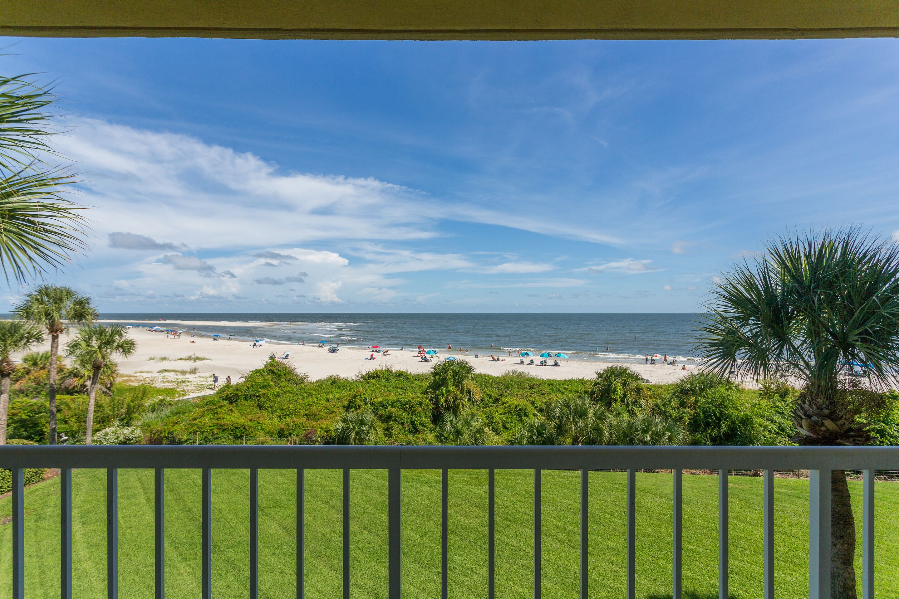 A beach view from a balcony with a railing, palm trees, and a clear sky.