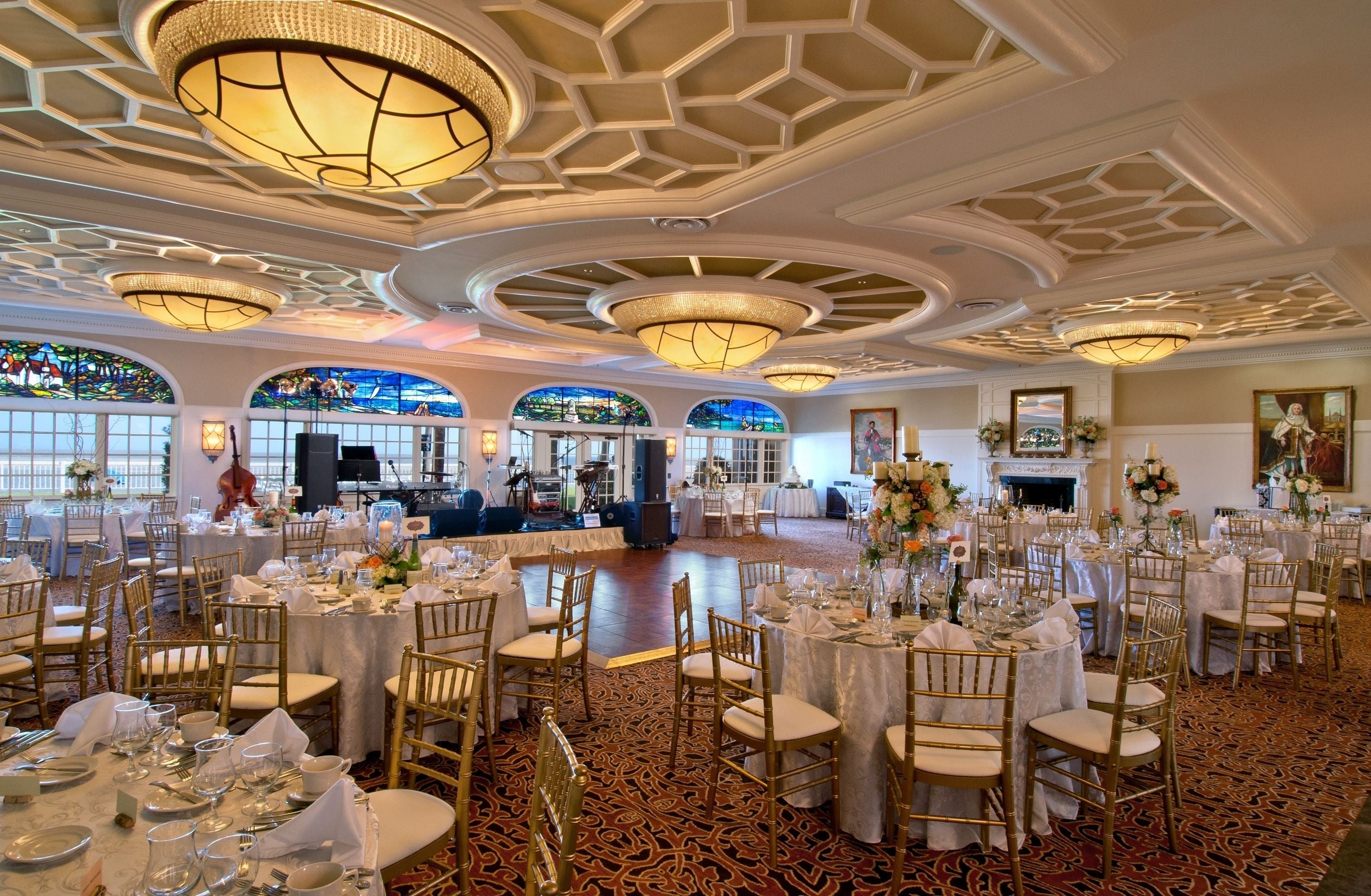 A banquet hall with round tables, gold chairs, and chandeliers.
