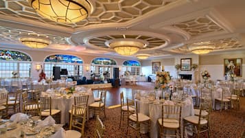A banquet hall with round tables, gold chairs, and chandeliers.