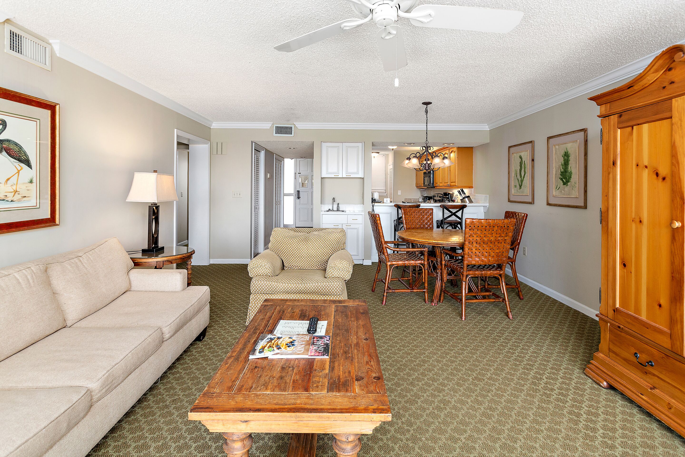 A living room with a sofa, coffee table, and dining area with chairs. There is a kitchen in the background.