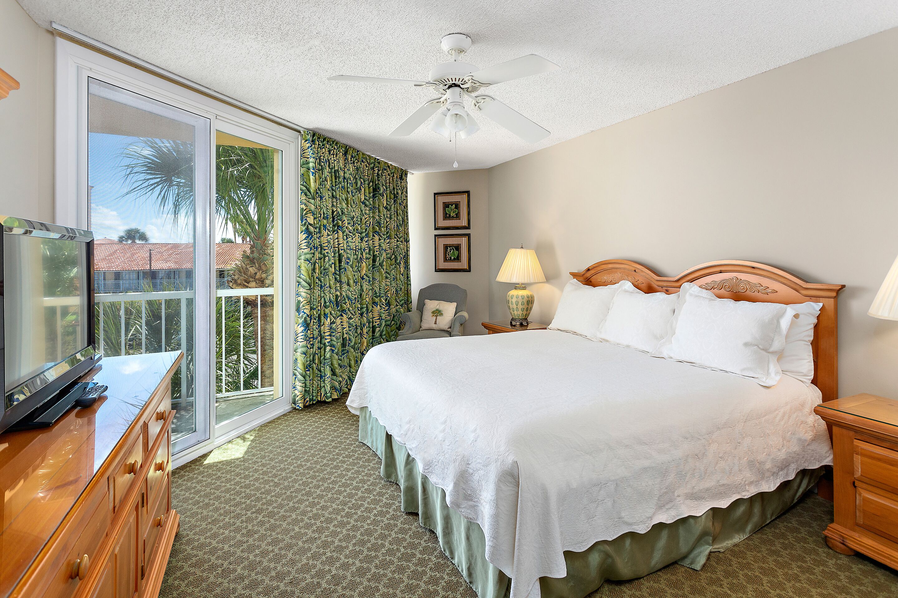 A hotel room with a bed, a television, a ceiling fan, a nightstand, a lamp, and a sliding glass door leading to a balcony.