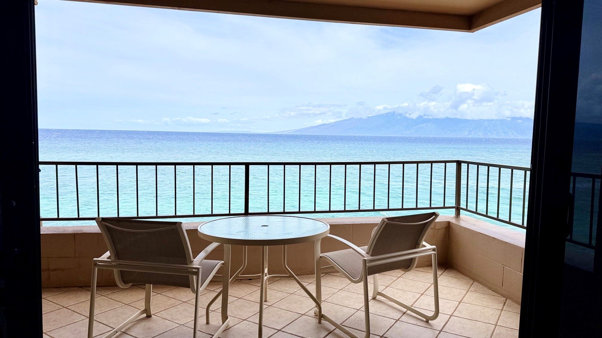 A balcony with two chairs and a table, overlooking a body of water and a distant mountain.