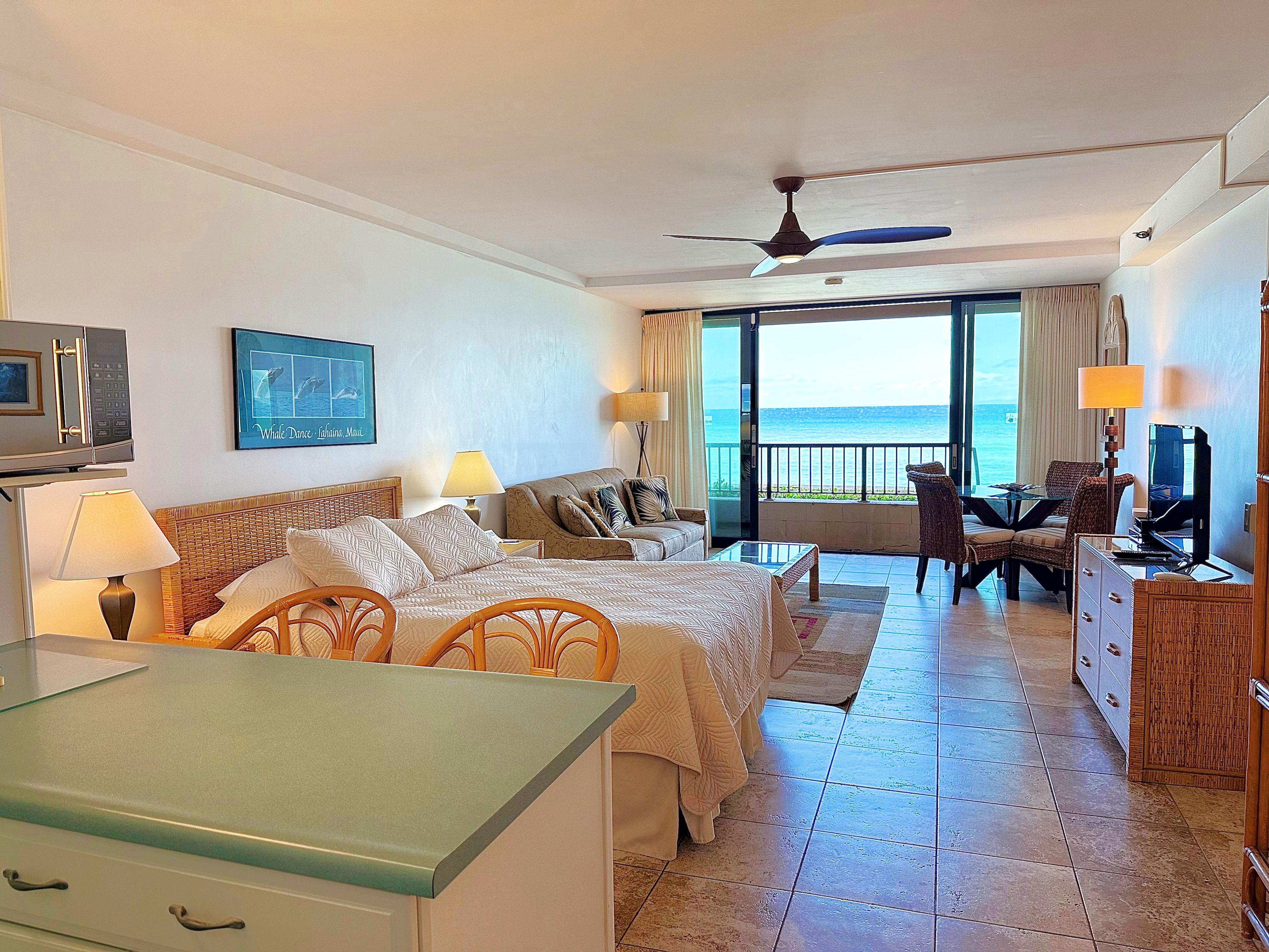 Ein Hotelzimmer mit Bett, Schreibtisch, Fernseher, einem Balkon mit Meerblick und einem Deckenventilator.