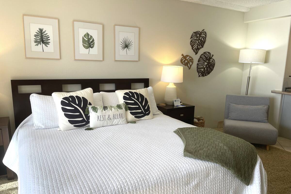 Una cama bien hecha con colcha blanca, varios almohadones, una mesita de noche con lámpara y decoración en la pared.