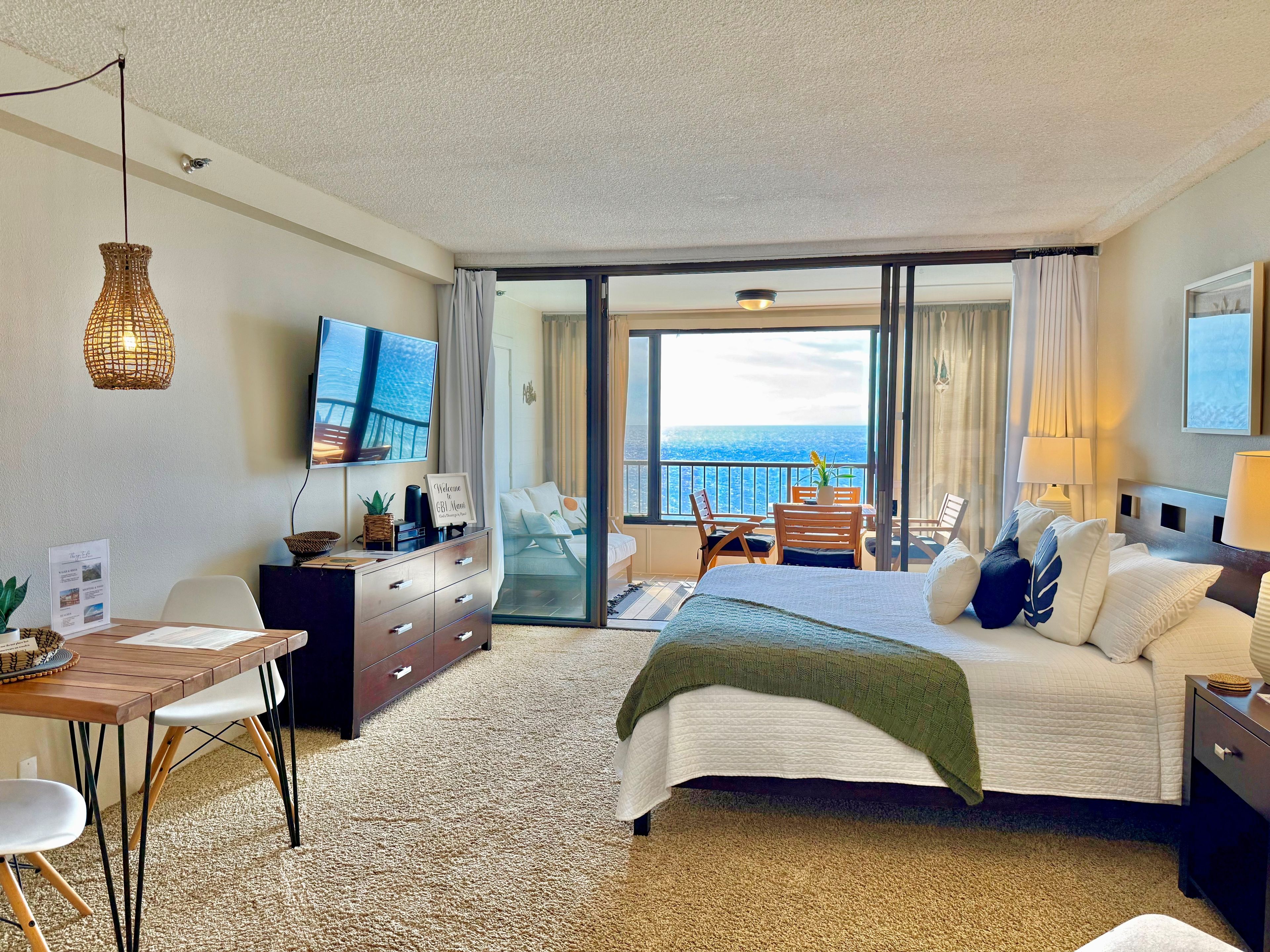 A hotel room with a bed, a desk, a TV, a balcony with a view of the sea, and a sliding door.