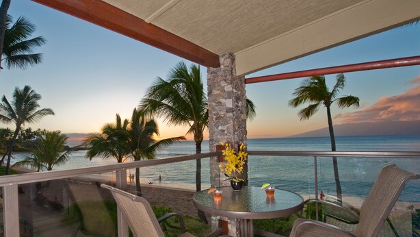 A balcony with a glass table, two wicker chairs, and a potted plant with yellow flowers. - The Mauian - Boutique Beach Studios on Napili Bay (Lahaina)