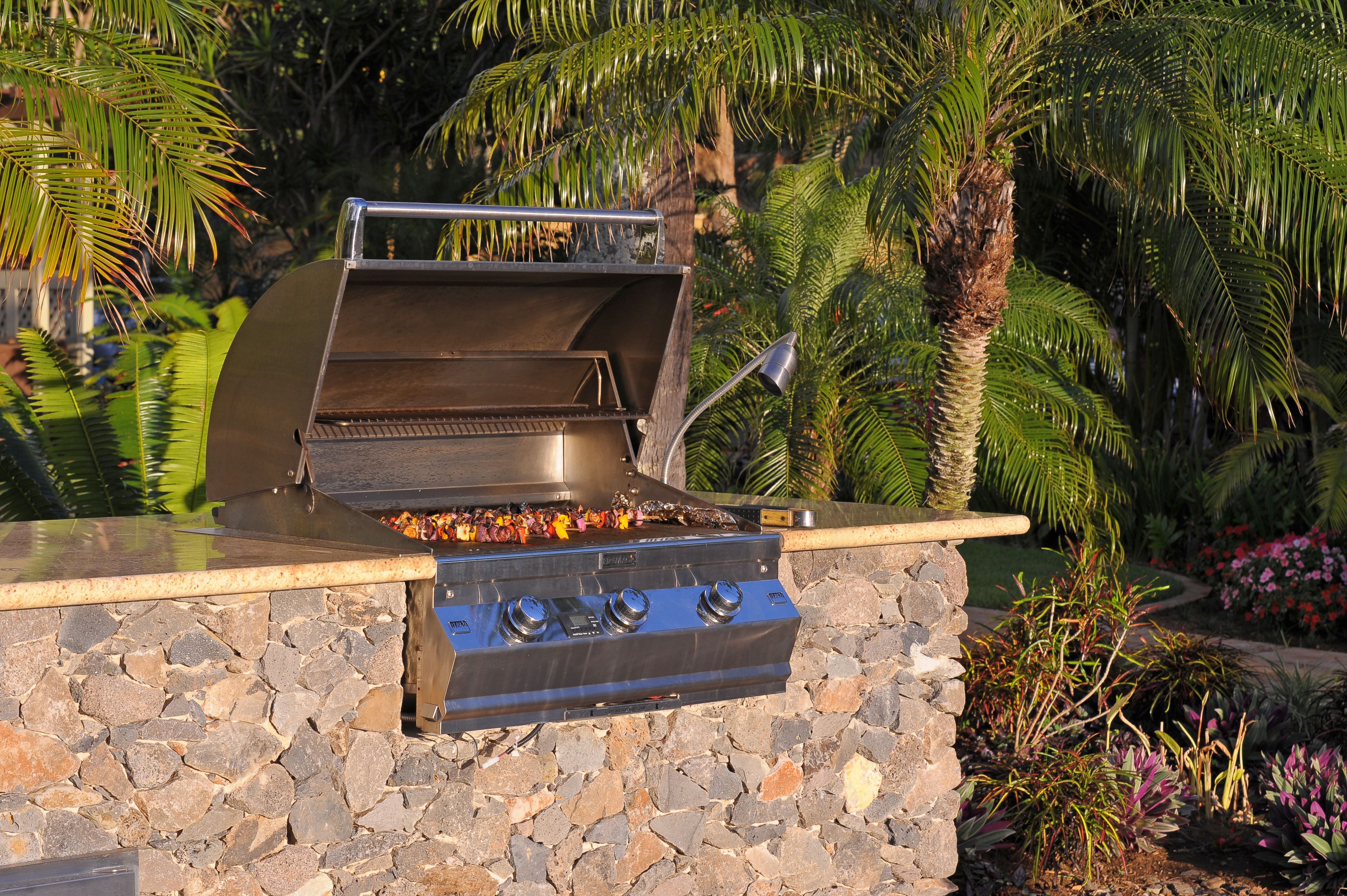 An outdoor grill with a stone base and a metal hood, surrounded by tropical plants and a palm tree.