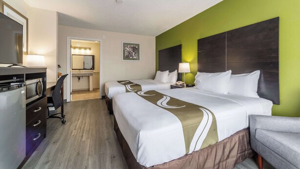 A hotel room with two beds, a desk, a chair, a TV, and a bathroom with a mirror and lights.