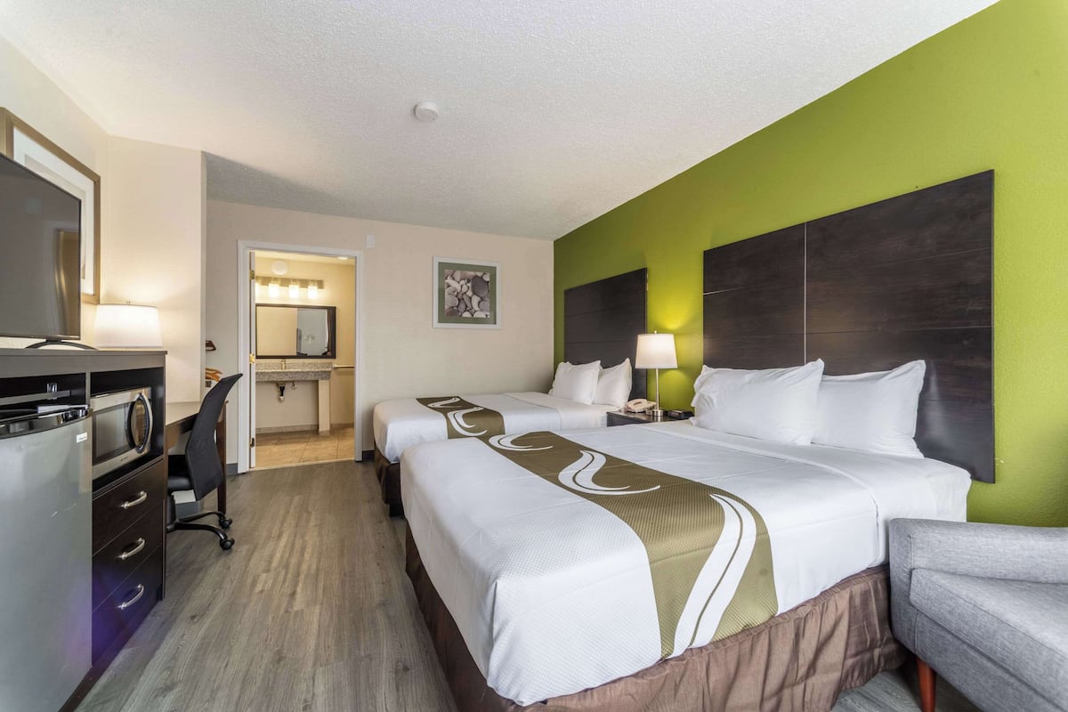 A hotel room with two beds, a desk, a chair, a TV, and a bathroom with a mirror and lights.