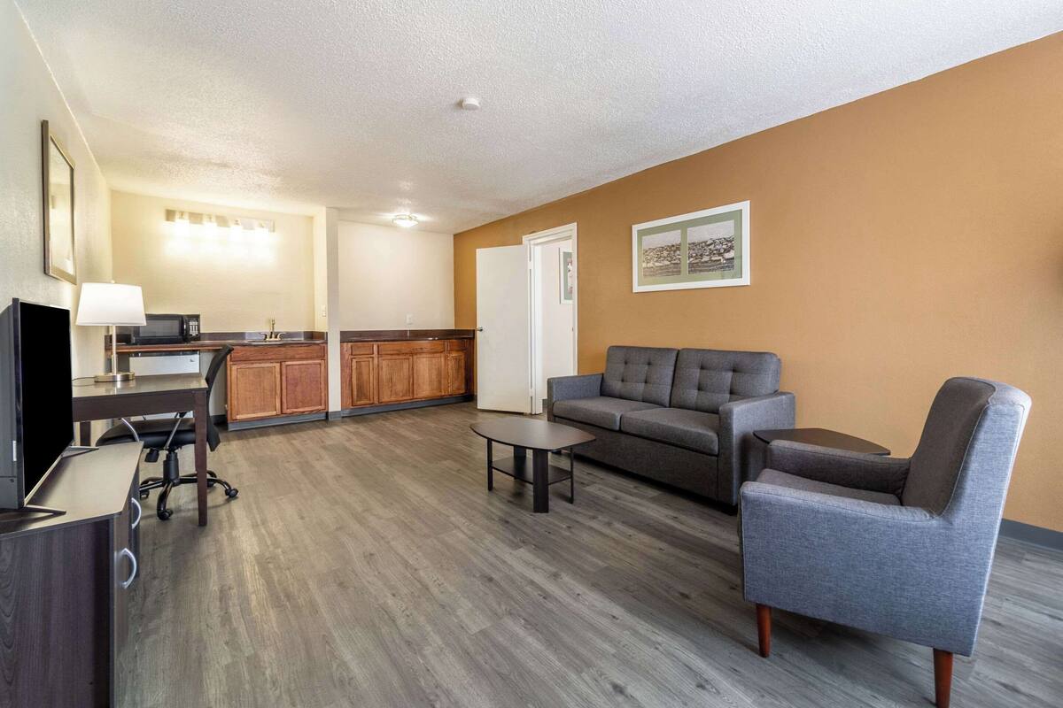 A modern living room with a grey sofa, a grey armchair, and a small coffee table. There is a TV on a wooden stand, a lamp, and a kitchen area with wooden cabinets and a microwave.