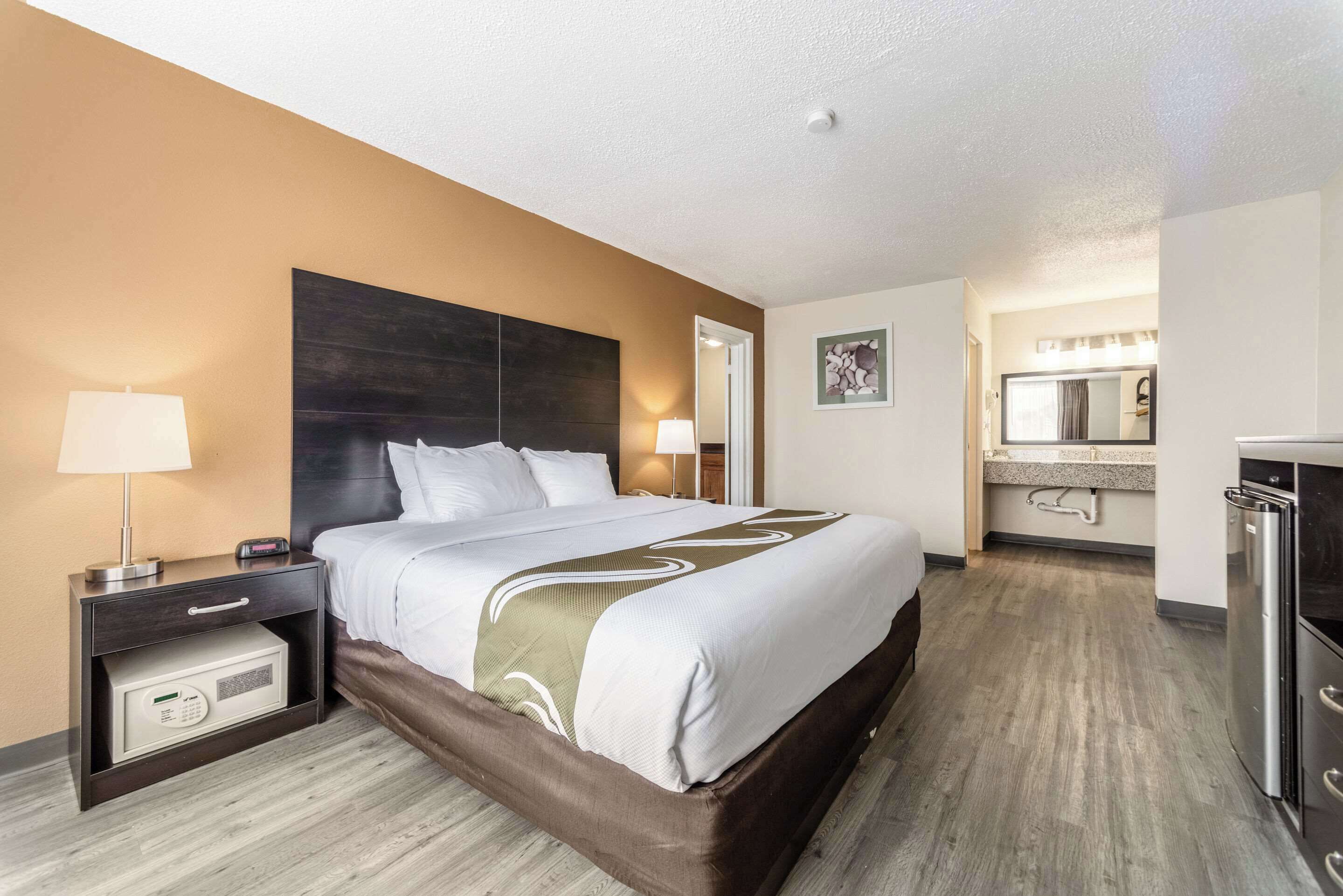 Habitación de hotel con una cama grande, mesitas de noche, microondas, refrigerador, baño con lavamanos y espejo, y piso de madera clara.
