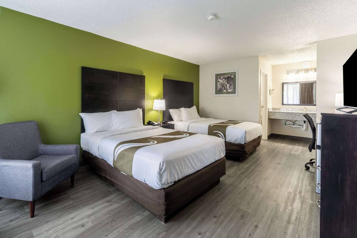 A hotel room with two beds, a gray armchair, a desk, and a bathroom visible through an open door.