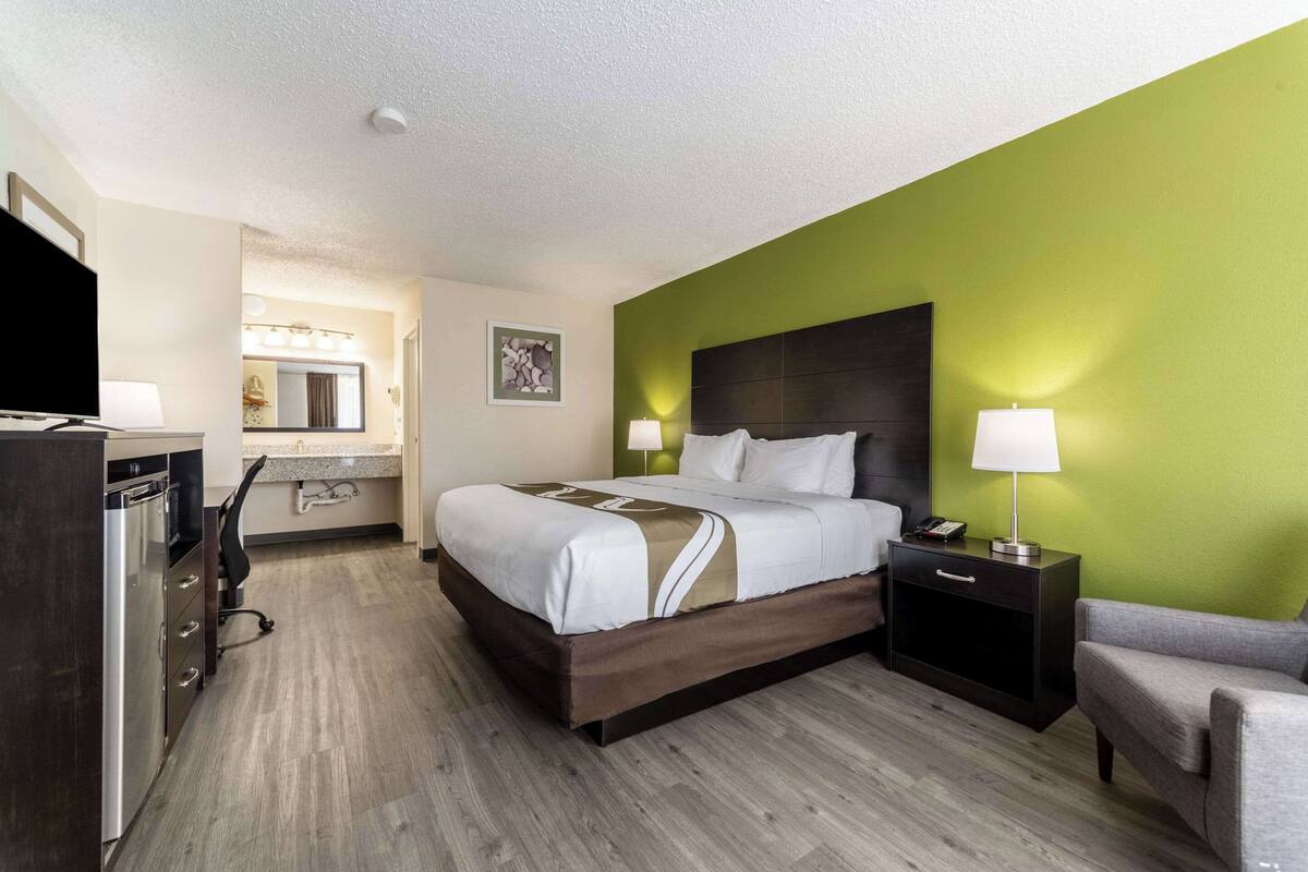 A hotel room with a bed, bedside table, chair, desk, and a television.