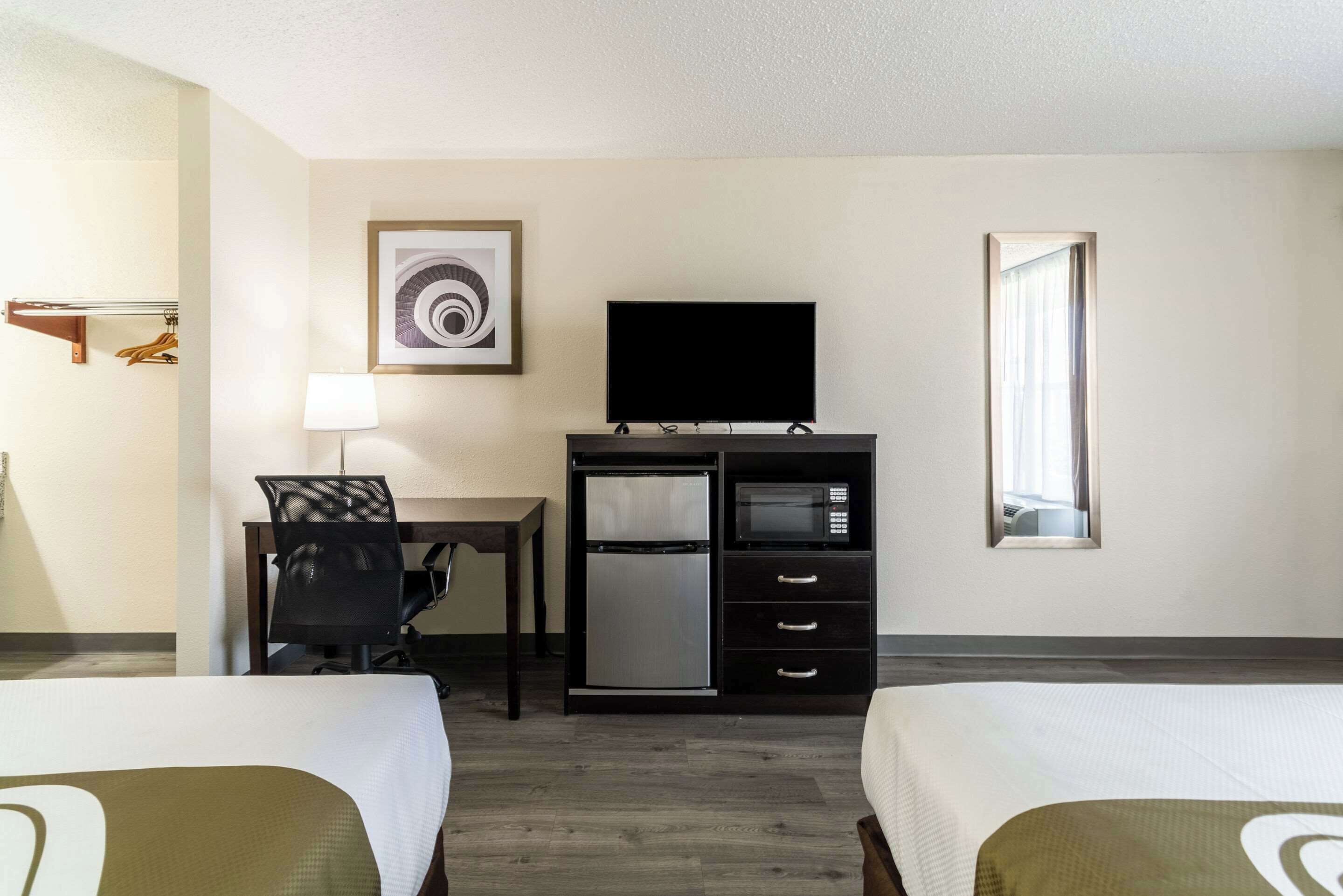 A hotel room with two beds, a desk with a chair, a flat-screen TV, a mini-fridge, a microwave, and a window with curtains.
