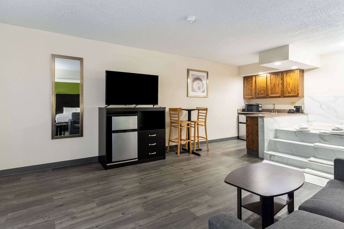 A modern living room with a flat-screen TV, a small kitchen with wooden cabinets, a dining area with two chairs, and a staircase leading to an upper level.