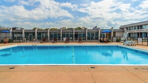 A swimming pool with lounge chairs and umbrellas, surrounded by a building with large windows and a flag.