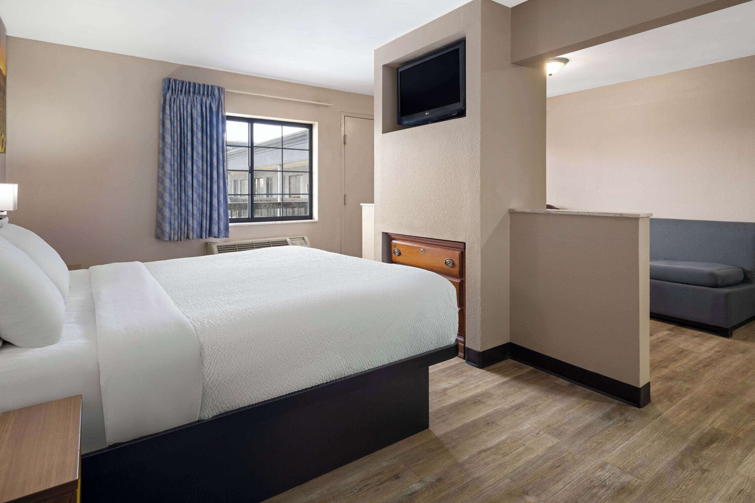 A hotel room with a bed, a TV mounted on the wall, a window with curtains, and a sofa.