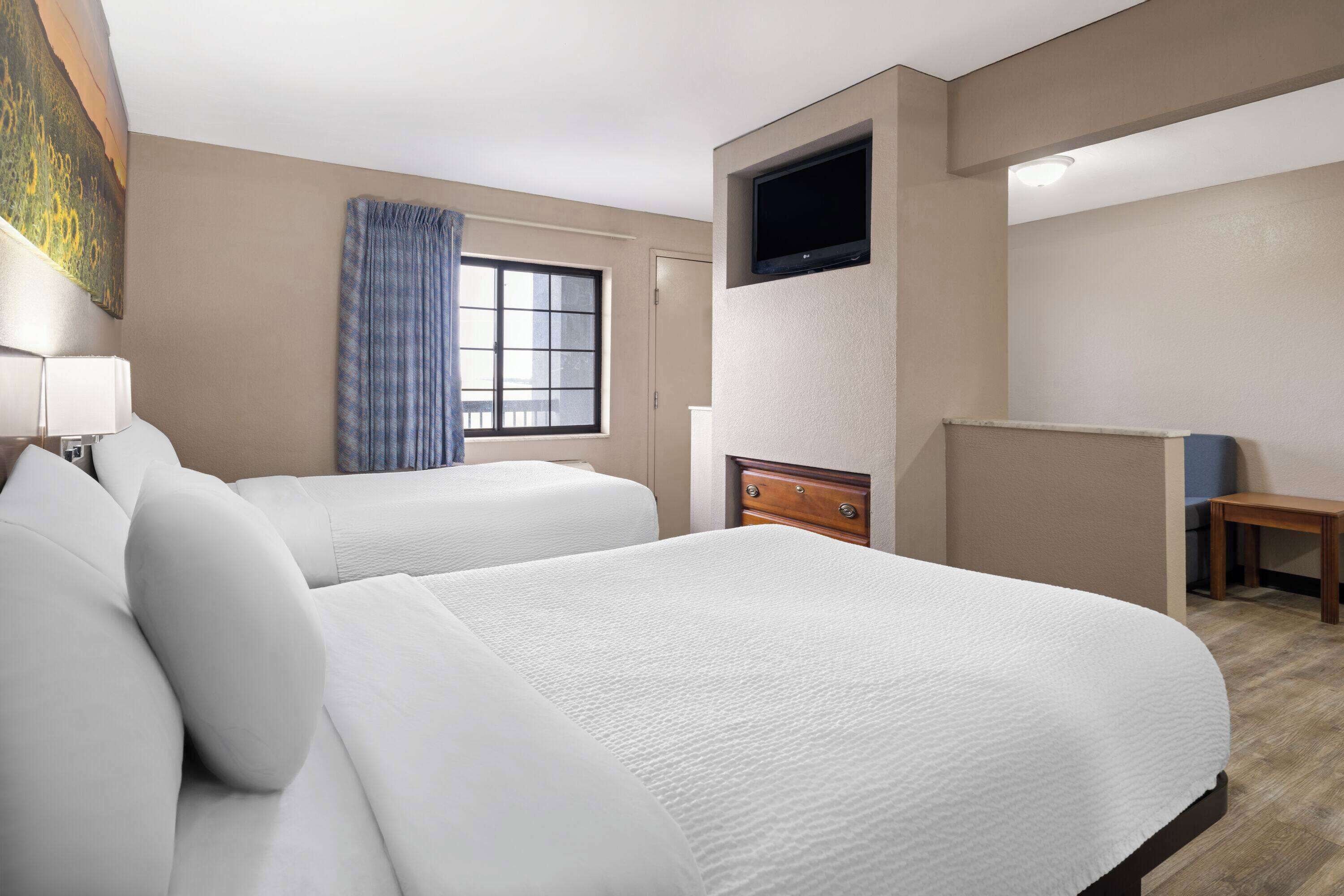 A hotel room with two beds, a television mounted on the wall, a window with curtains, and a small table.
