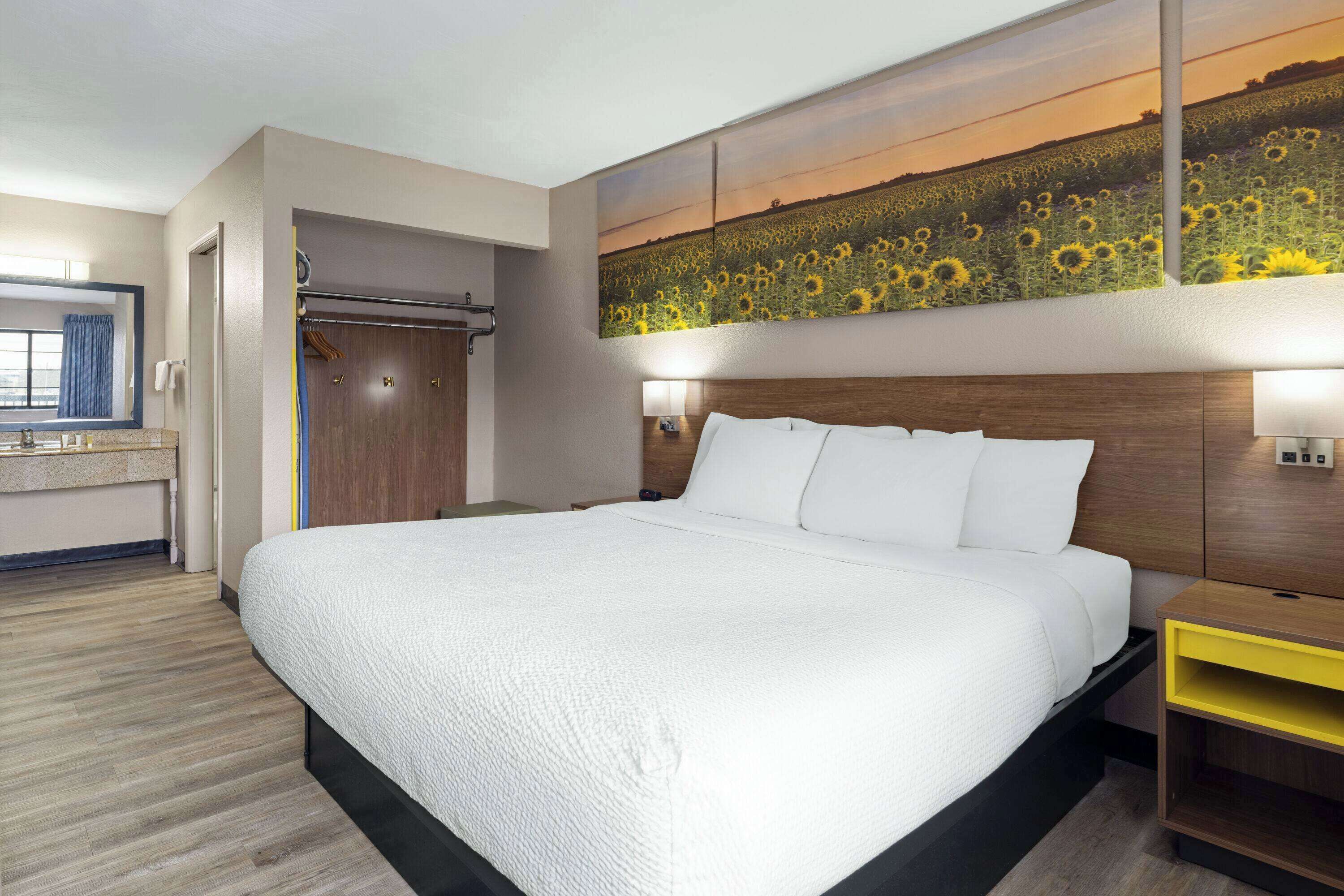 Une chambre d’hôtel avec un grand lit, une tête de lit en bois, une table de chevet, une lampe fixée au mur et un tableau représentant des tournesols.
