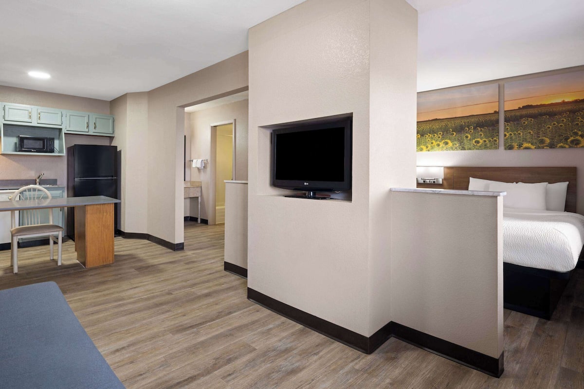 A hotel room with a kitchenette, a flat-screen TV, a bed, and a painting of sunflowers.