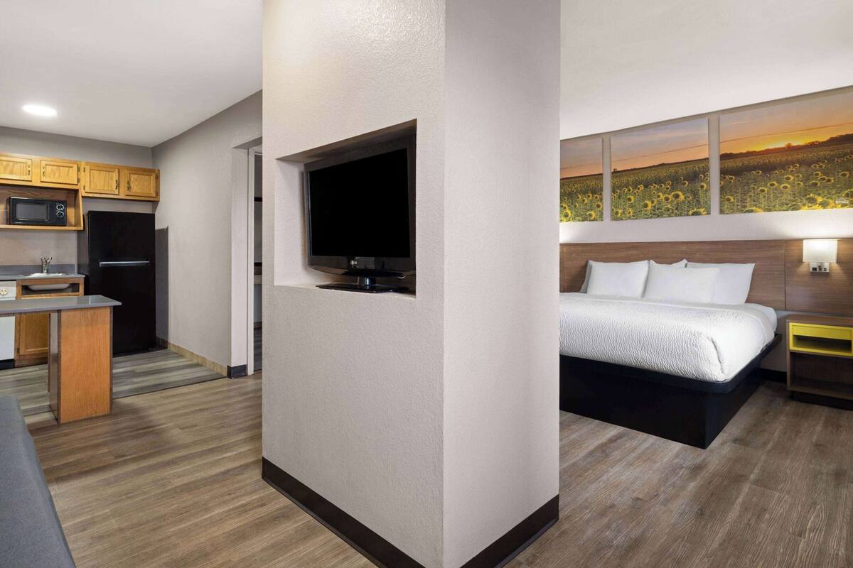 A compact hotel room with a kitchenette, a flat-screen TV, a bed with white bedding, and a wall-mounted artwork depicting a sunset over a field.