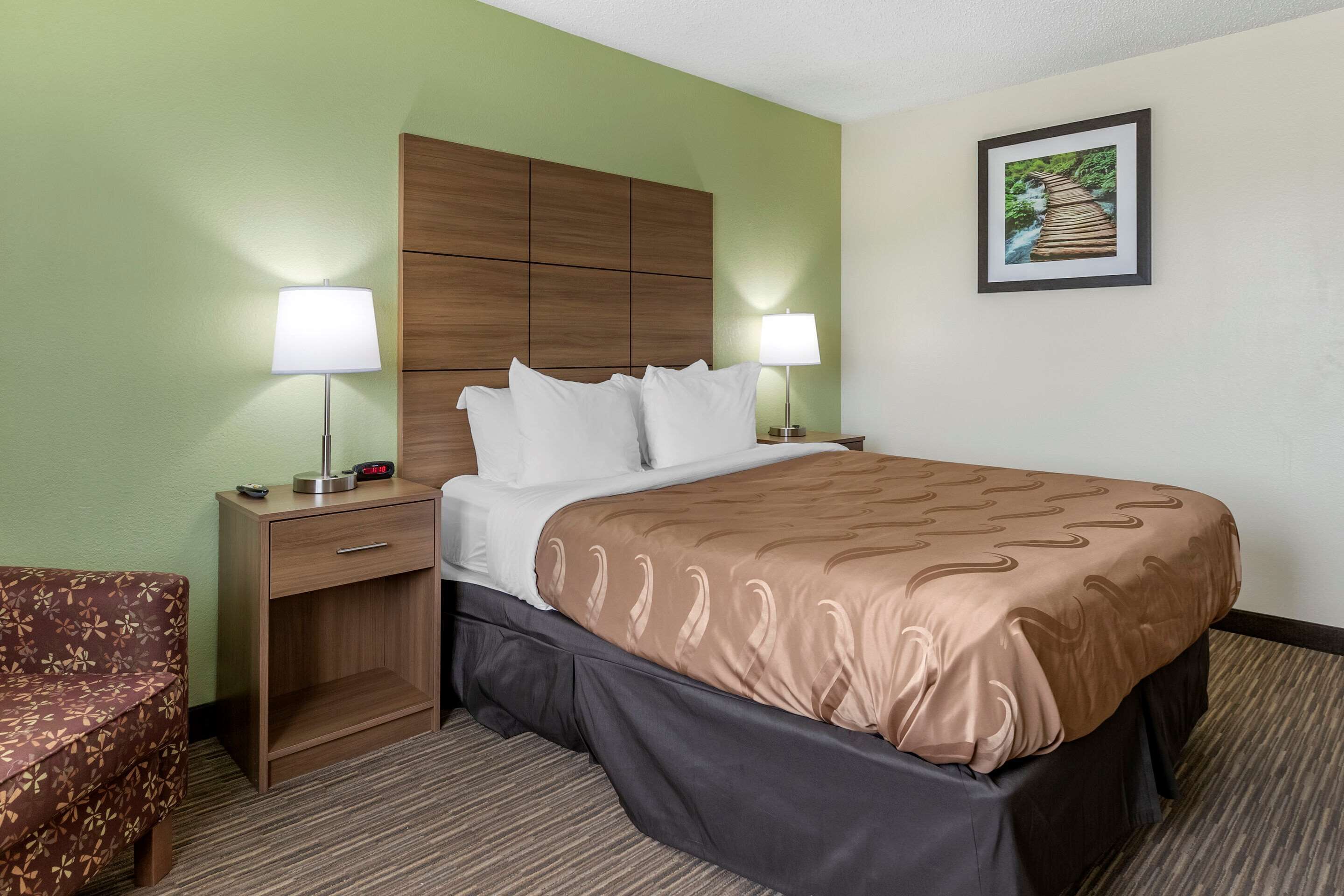 A hotel room with a bed, two bedside lamps, a nightstand, a wooden headboard, a framed picture on the wall, and a patterned sofa.