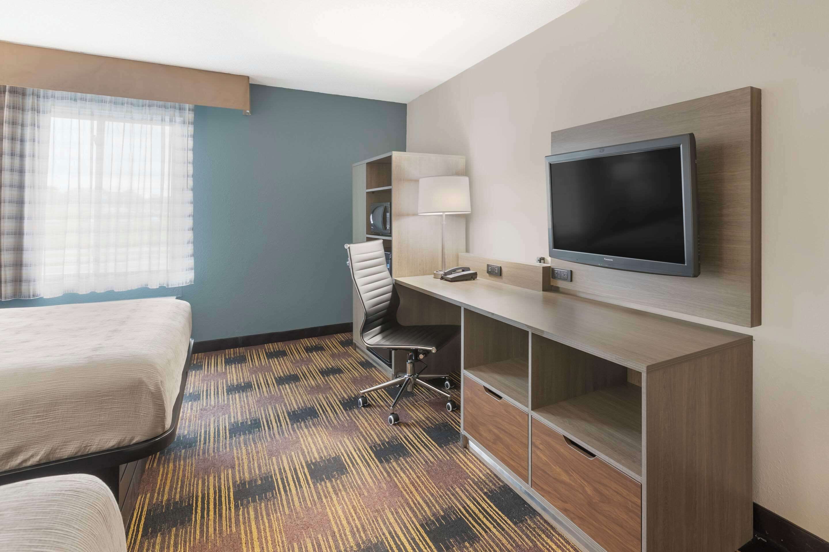 A hotel room with a bed, a desk with a flat-screen TV, a chair, and a lamp.