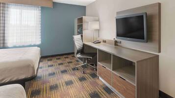 A hotel room with a bed, a desk with a flat-screen TV, a chair, and a lamp.