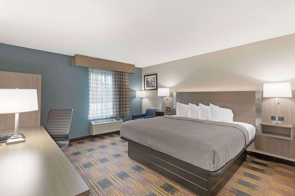 A hotel room with a large bed, a desk with a lamp, a chair, a window with curtains, and a picture on the wall.