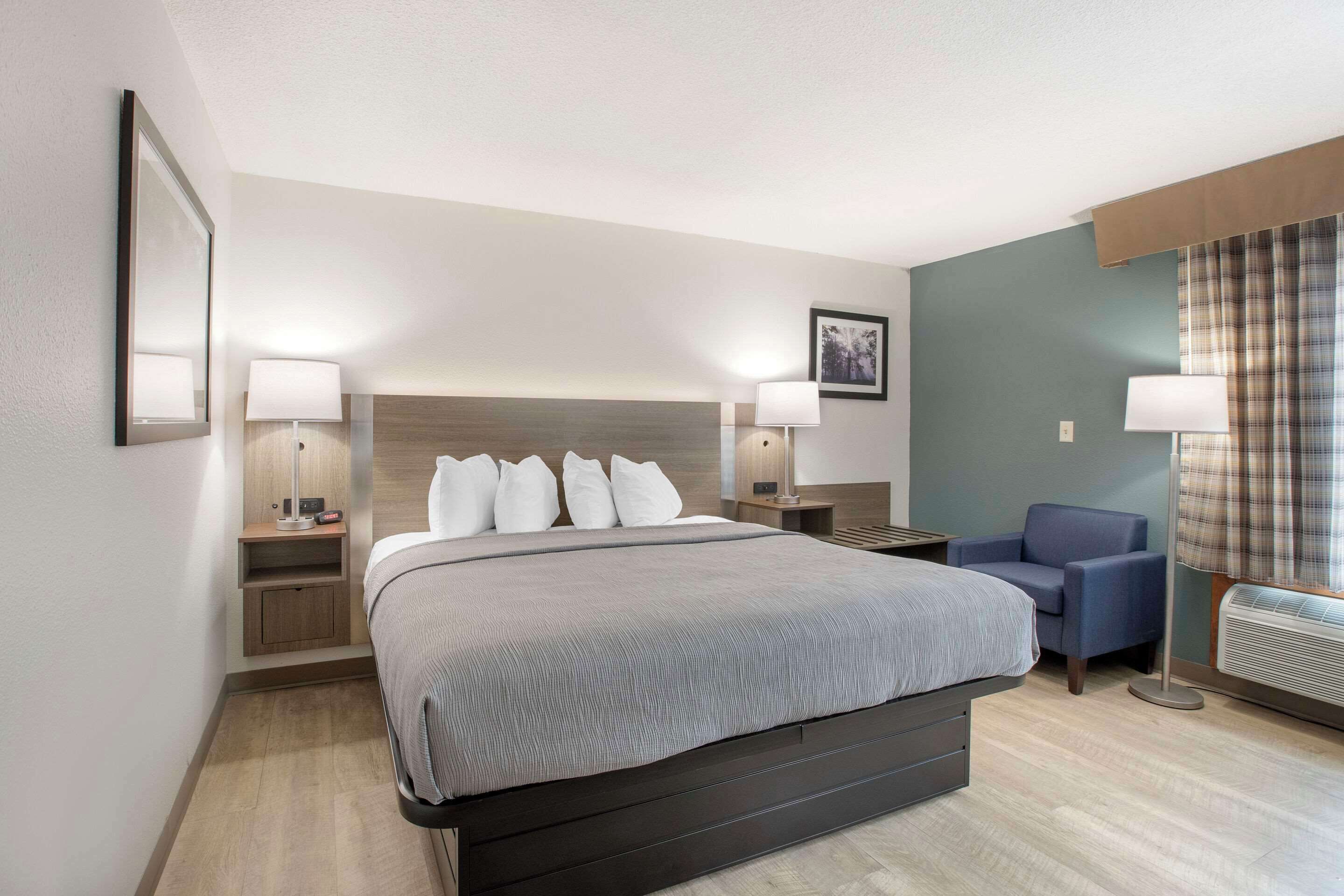 Una habitació d'hotel amb un llit gran, dues taules de nit amb làmpades, una cadira, un quadre a la paret i una finestra amb cortines.
