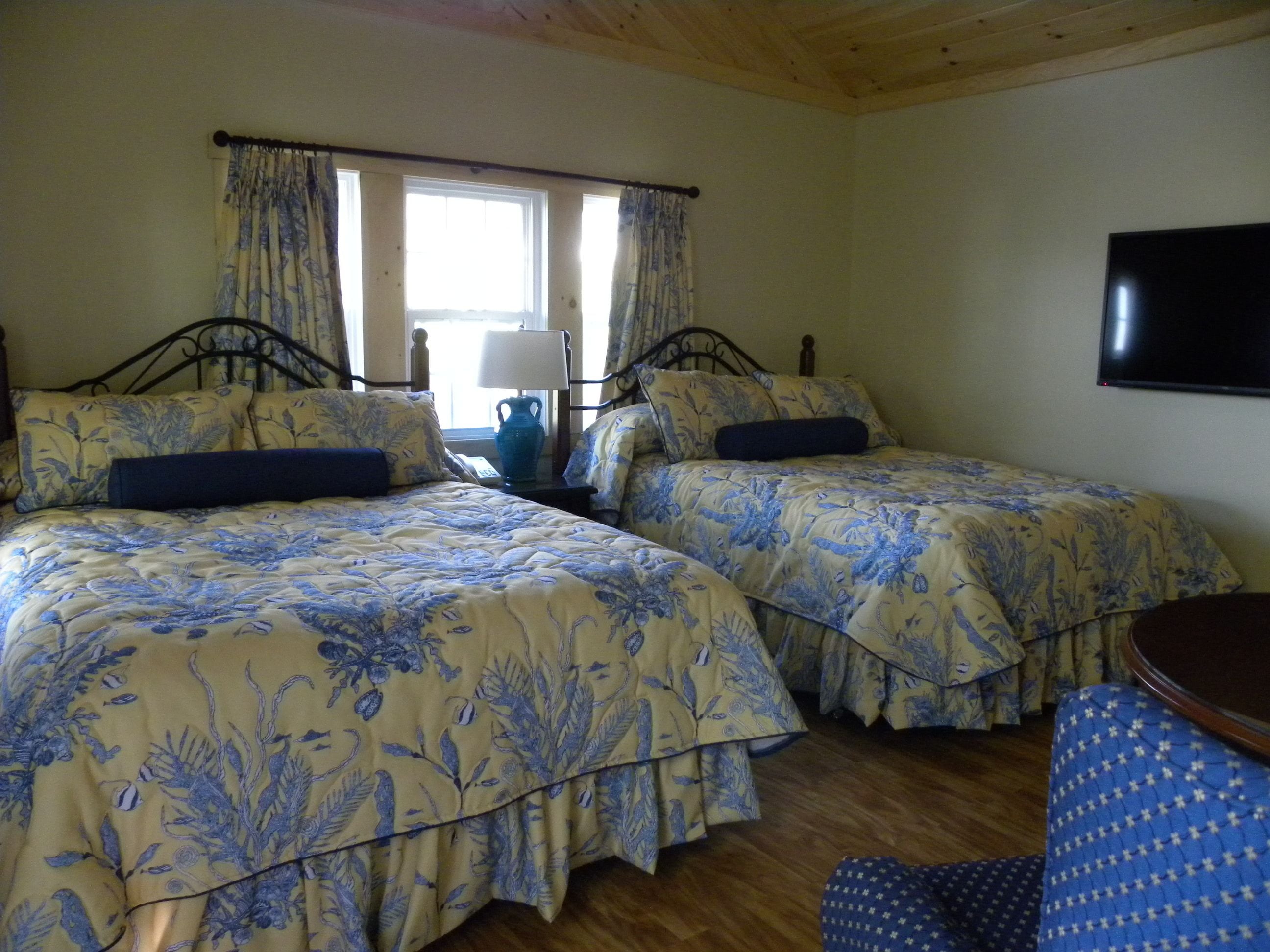 A room with two beds, a wooden ceiling, a window with curtains, and a flat-screen TV on the wall.