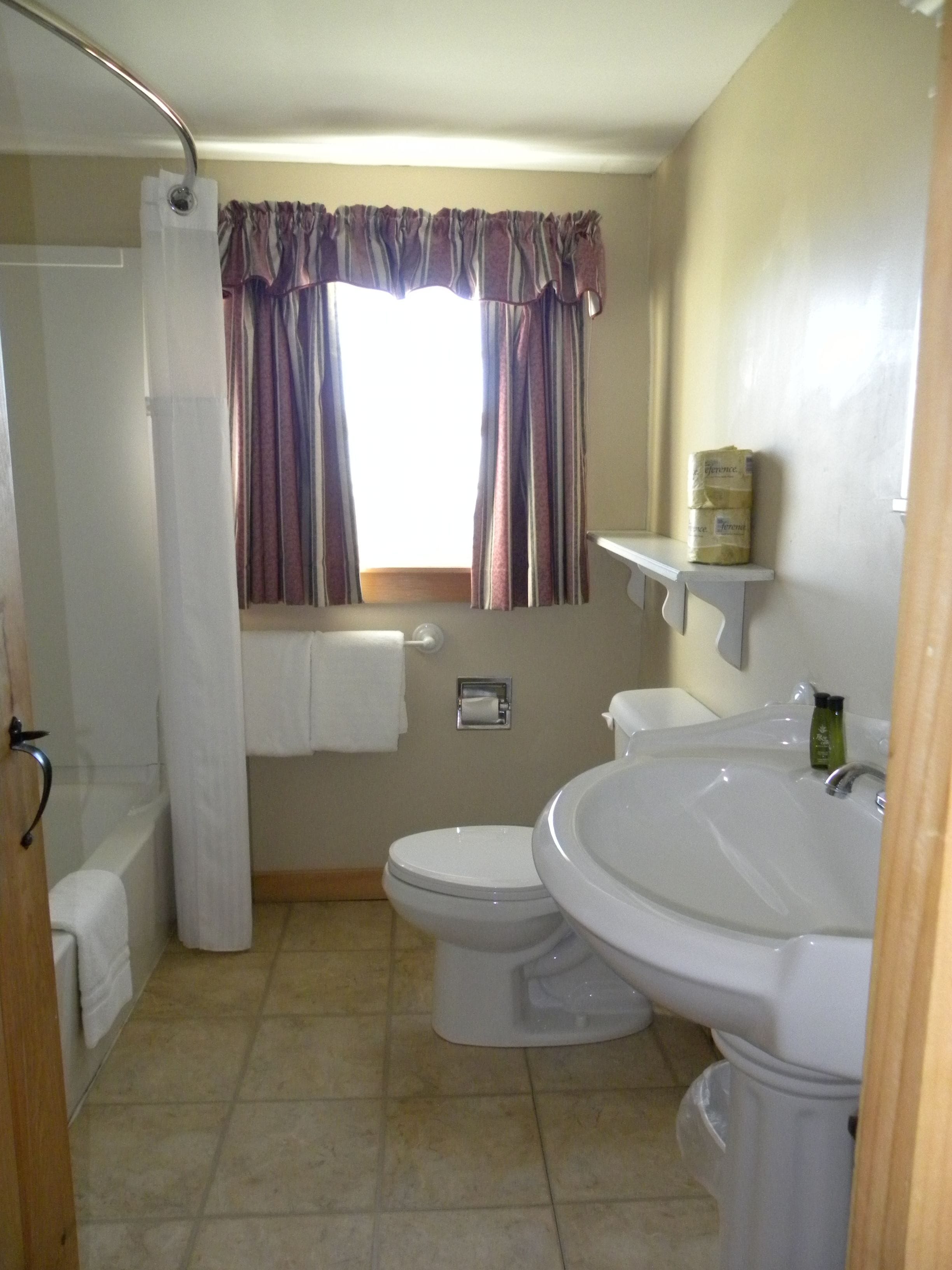 A bathroom with a toilet, bidet, and sink. There is a window with curtains, a towel rack, and a toilet paper holder.